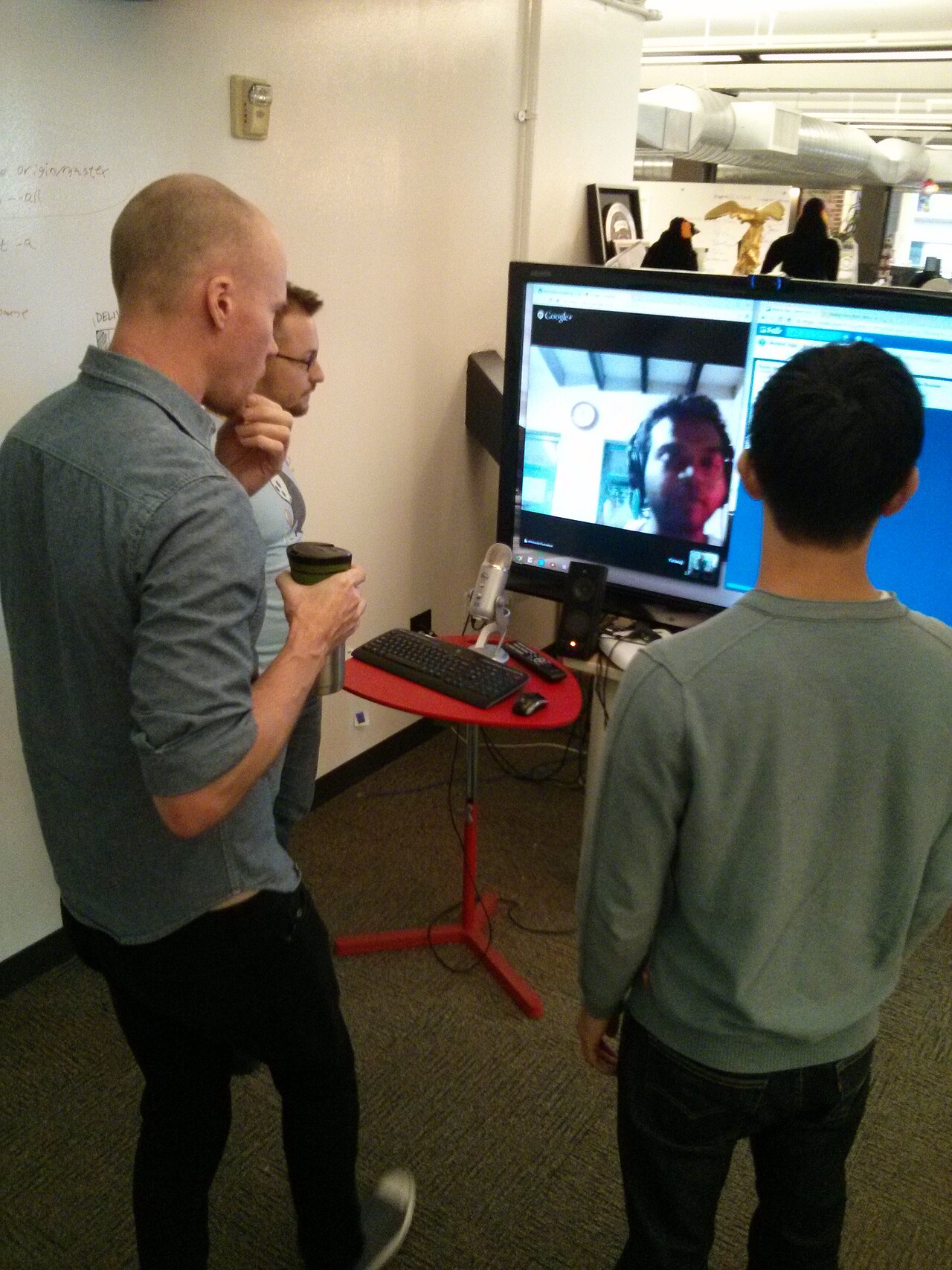 Three people standing at a screen during a team stand-up meeting, with a remote colleague joining by video call and a microphone on a stand beside the display